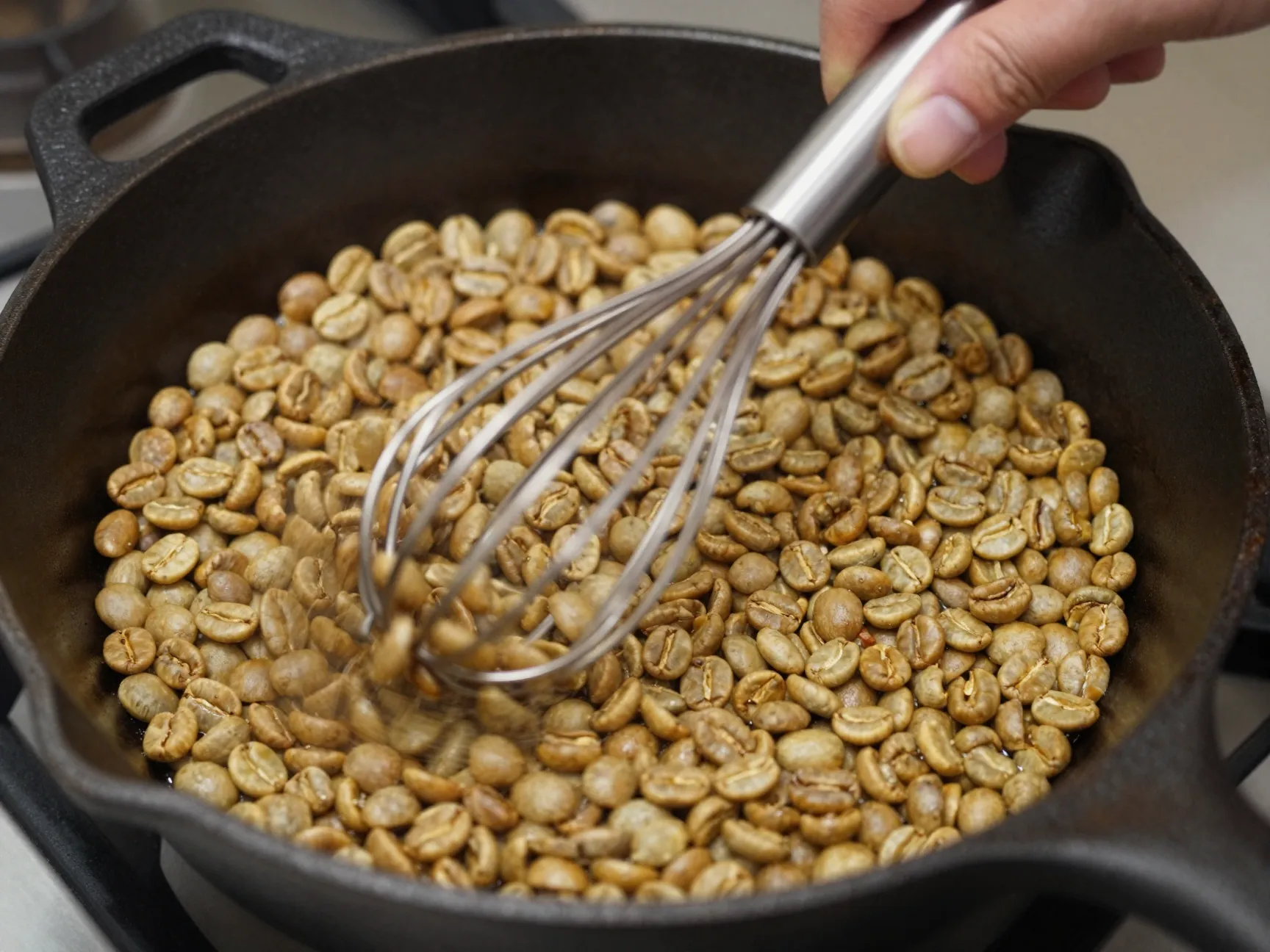Hand stirring coffee beans constantly with wire whisk in cast iron skillet