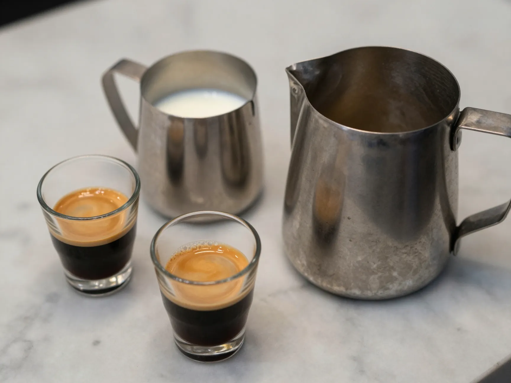 Two espresso shots beside small milk pitcher and large milk pitcher